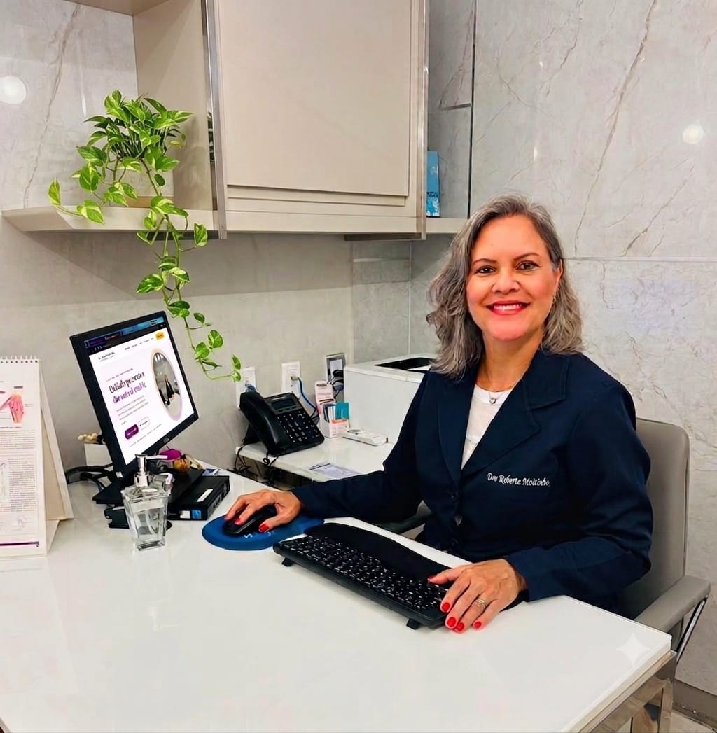 Dra. Roberta Leal sorridente, sentada à mesa de seu consultório com jaleco azul-marinho